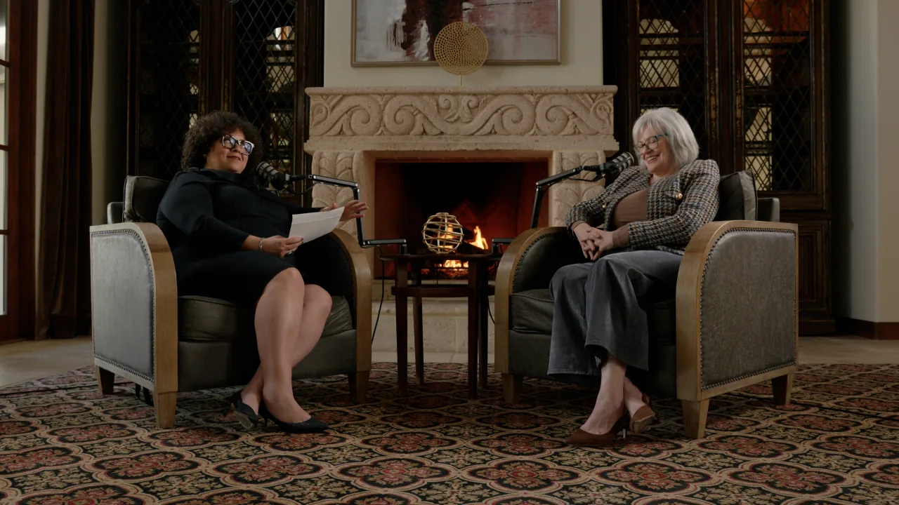 Two people recording an interview in armchairs beside a fireplace.